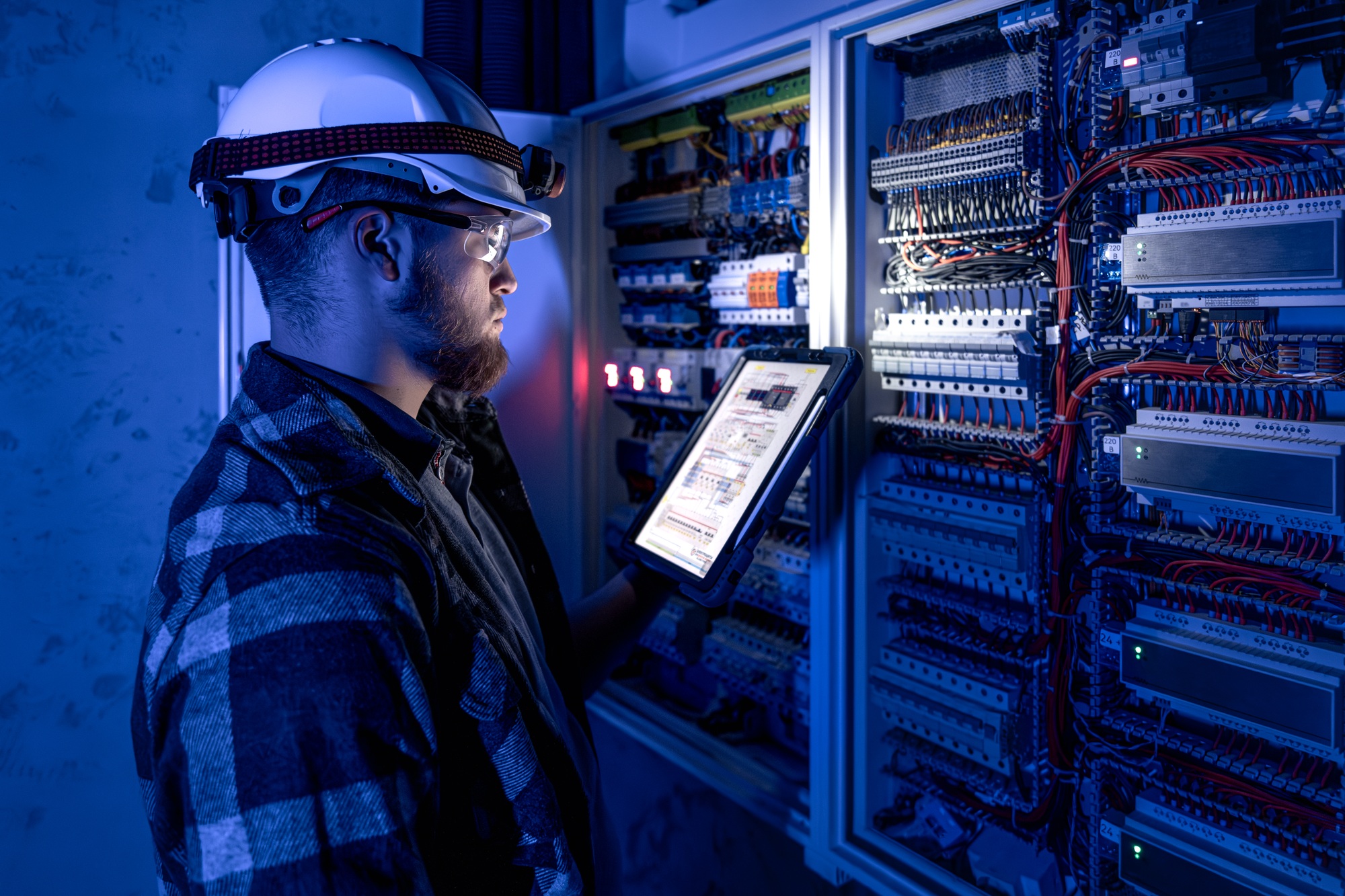 Electrician in overalls, focused on work in switchboard with fuses, using tablet.